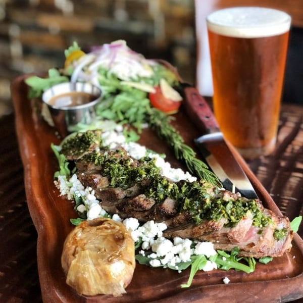 Grilled steak with chimichurri, feta, roasted garlic, salad, and a beer on a wooden board.