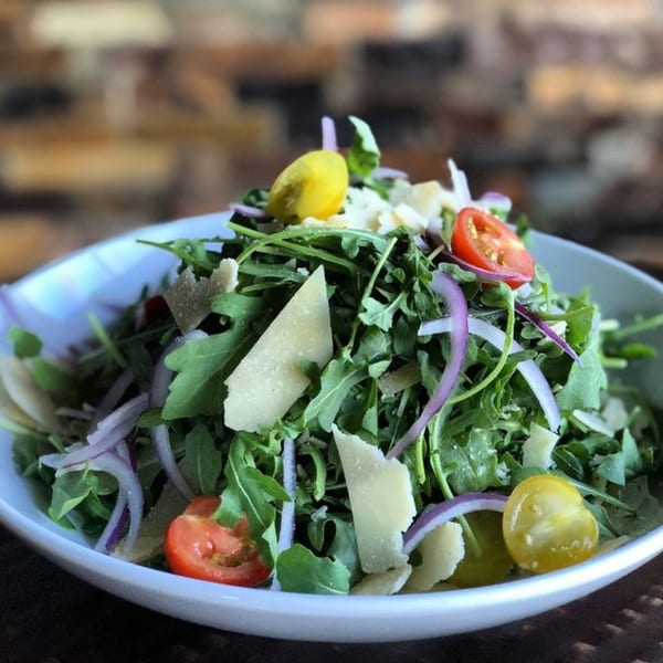 A fresh arugula salad with shaved parmesan, red onion, and cherry tomatoes in a white bowl.