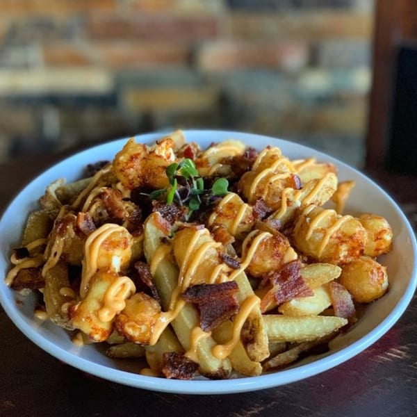 A white bowl of golden fries and tater tots topped with bacon bits and a creamy orange sauce.